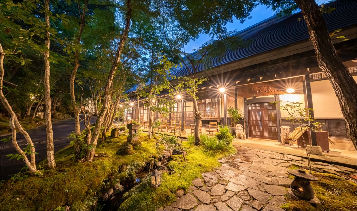 Illuminated wooden entrance and stone walkway of the Yufuin ryokan at dusk surrounded by moss and trees