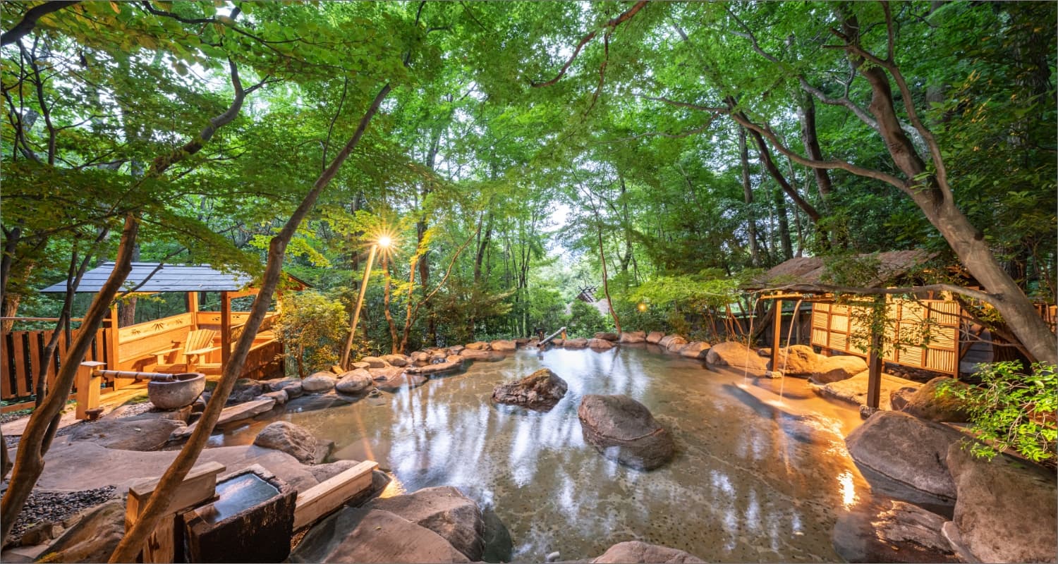Forest rotenburo outdoor onsen with rocks and thatched bath huts amid greenery in Yufuin
