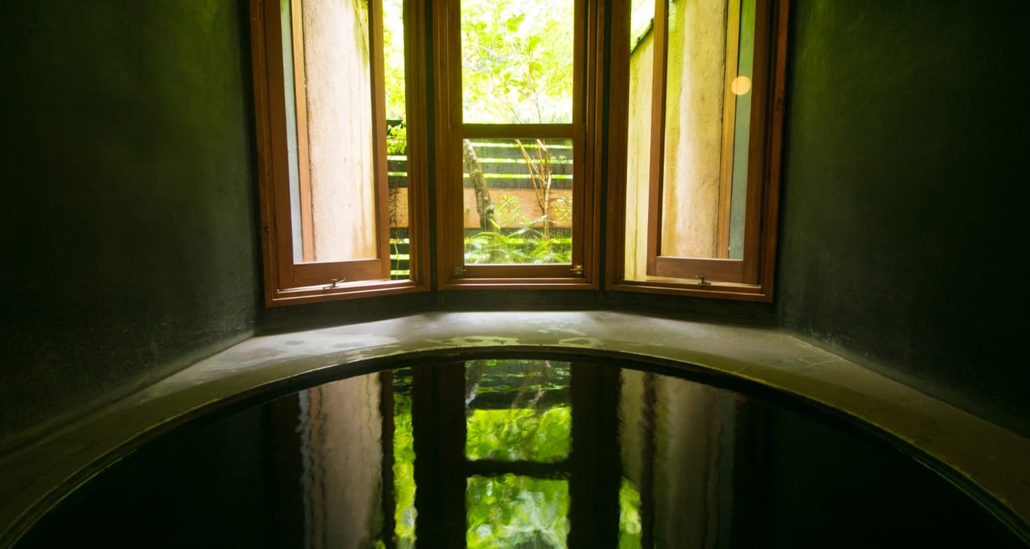 Indoor stone soaking bath with green clear water and a wooden window opening onto the garden