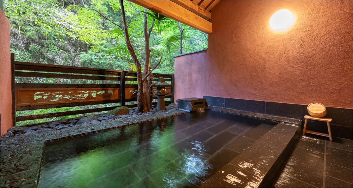 Private black-stone rotenburo open-air bath beside a maple tree and wooden fence in Yufuin