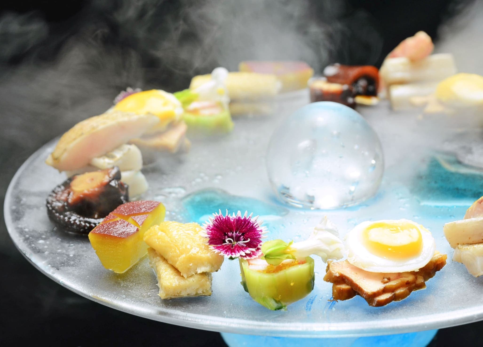 Hatago kaiseki appetizer platter of seasonal morsels arranged on a glass plate amid dry-ice mist