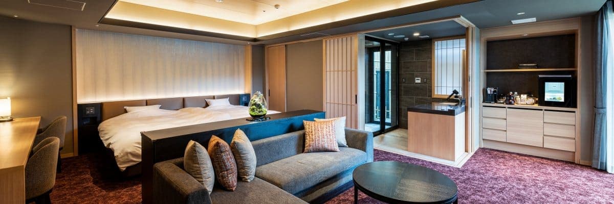 Hatago Room 101 suite with a low bed, grey sofa and an en-suite private bath visible behind sliding shoji doors