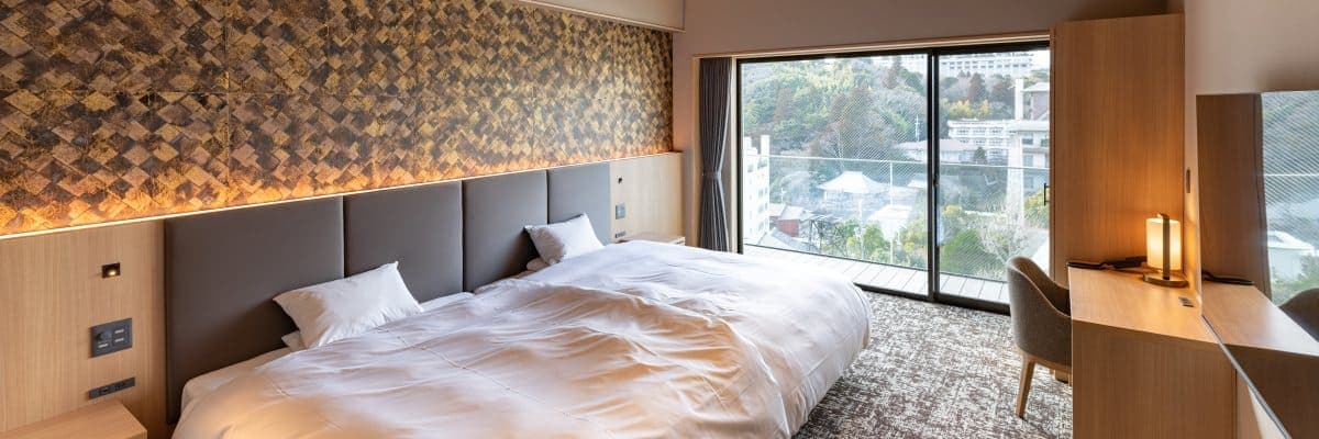 Hatago Room 302 bedroom with a wide bed beneath a gold mosaic headboard wall and a balcony view of the hills