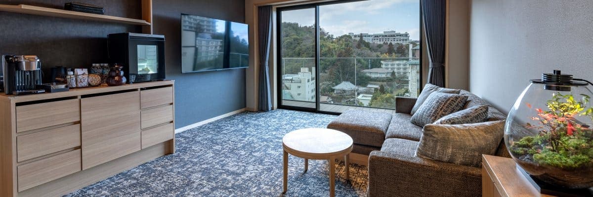 Hatago Room 301 living area with a corner sofa, low round table and floor-to-ceiling window over the Arima townscape