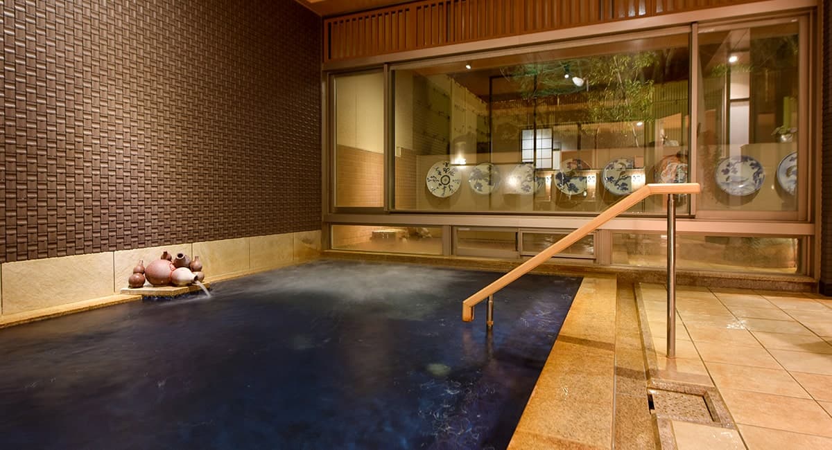 Indoor bath at Oku no Hosomichi with clear ginsen spring water, tiled walls and a row of decorative ceramic plates lit at dusk