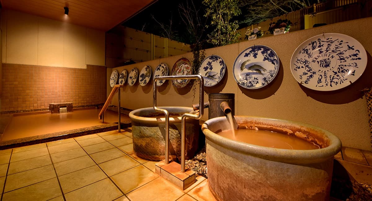 Open-air kinsen rotenburo at Oku no Hosomichi with ceramic tubs of reddish Arima gold spring water below a wall of blue-and-white Imari plates