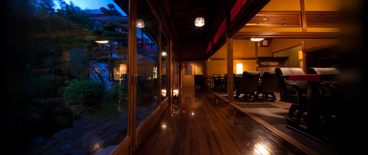 Lantern-lit wooden corridor and lounge with leather chairs reflecting the night garden through glass, Ryokan Sennari, Beppu Onsen