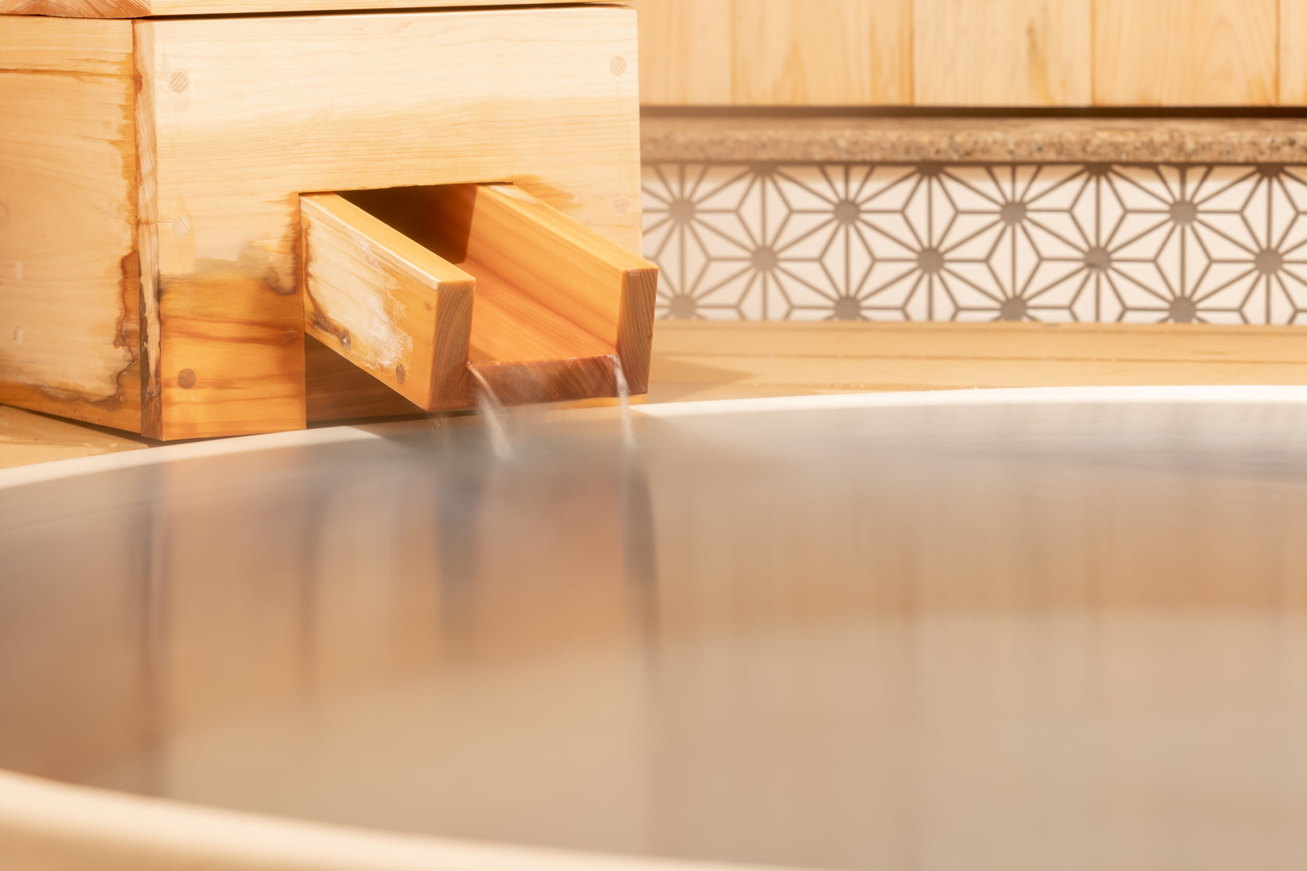Wooden spout pouring spring water into a round bath beside an asanoha-pattern tile border
