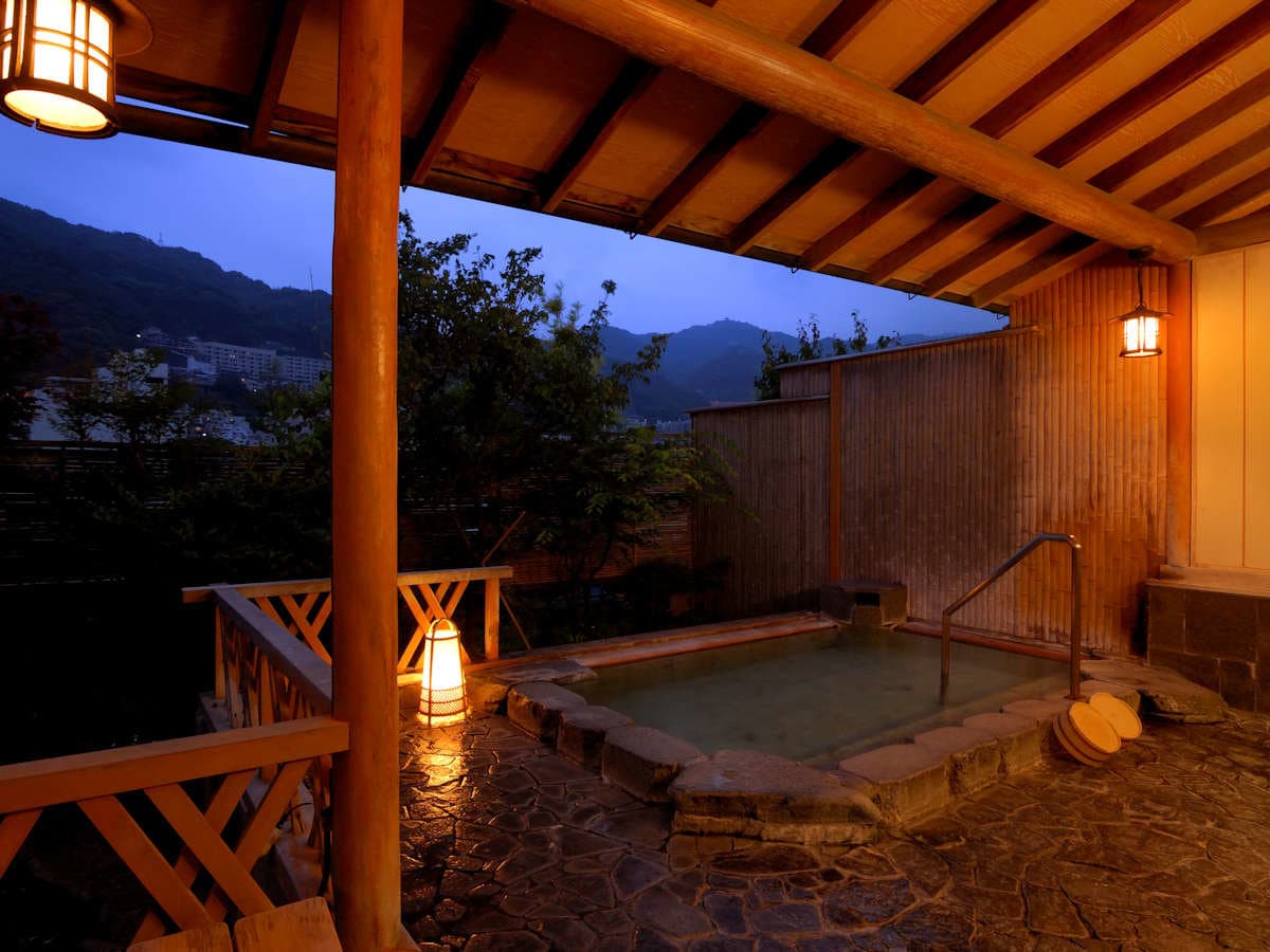 Rooftop open-air rotenburo at dusk with stone surround, lanterns and mountain views in Atami Onsen