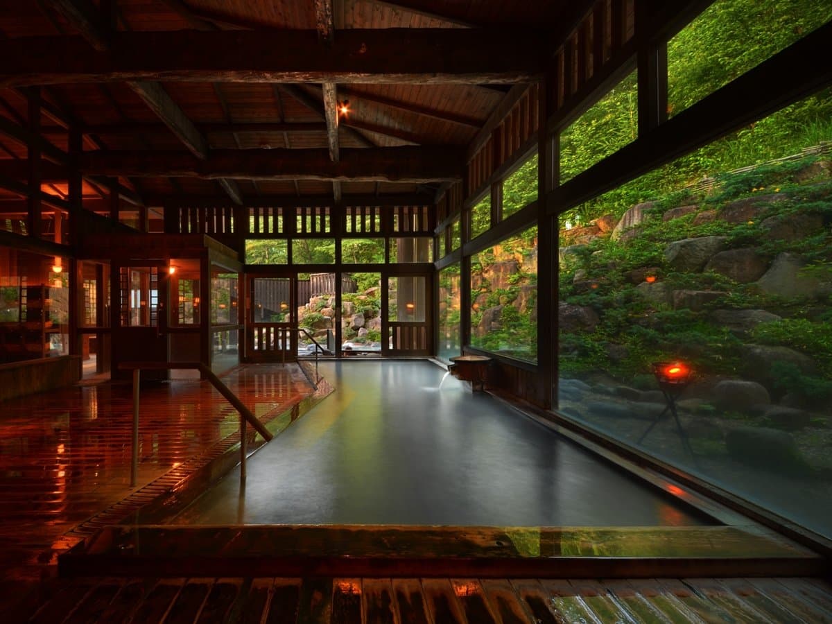 Timber-framed indoor bathhouse at dusk with warm lanterns and a garden view at Zao Kokusai Hotel