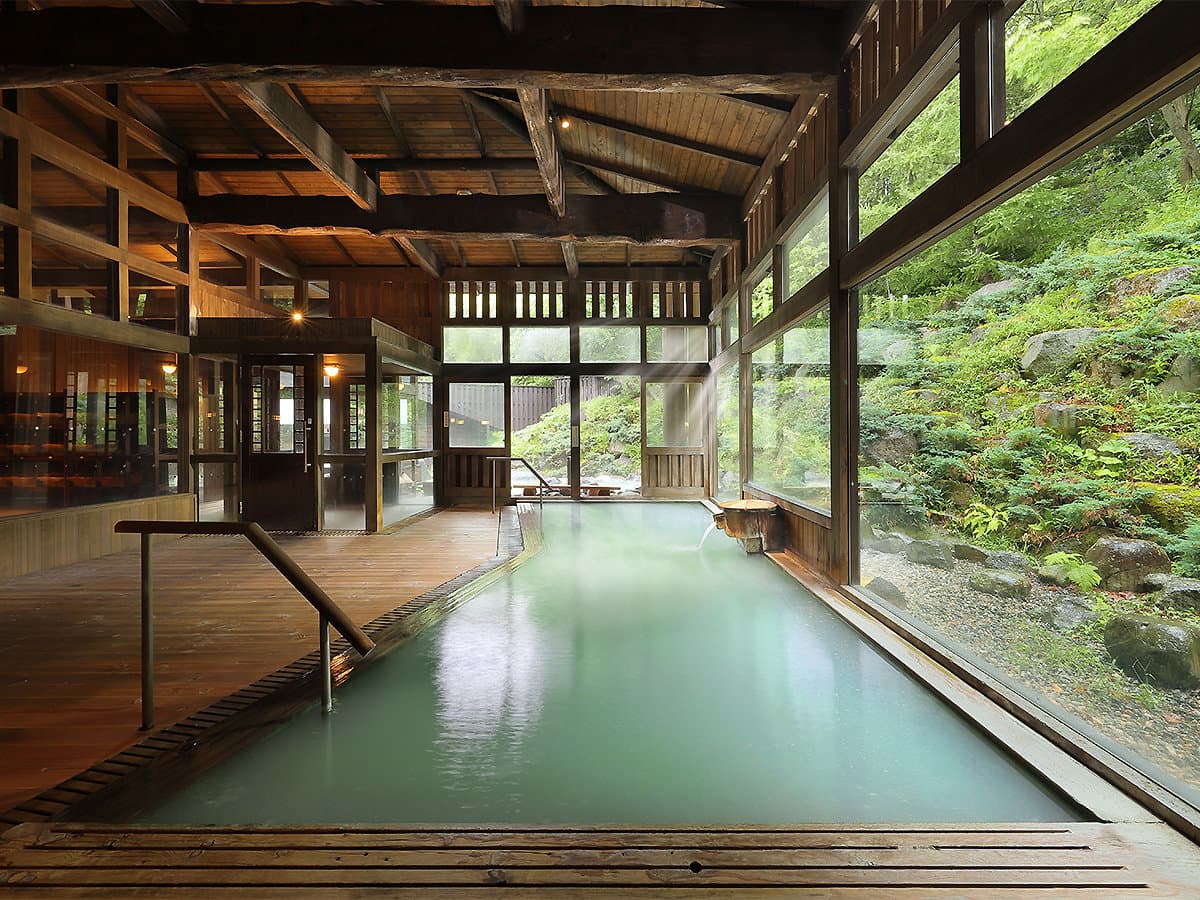 Spacious wooden indoor onsen bath with sulfur-clouded water and a wall of windows facing the forest