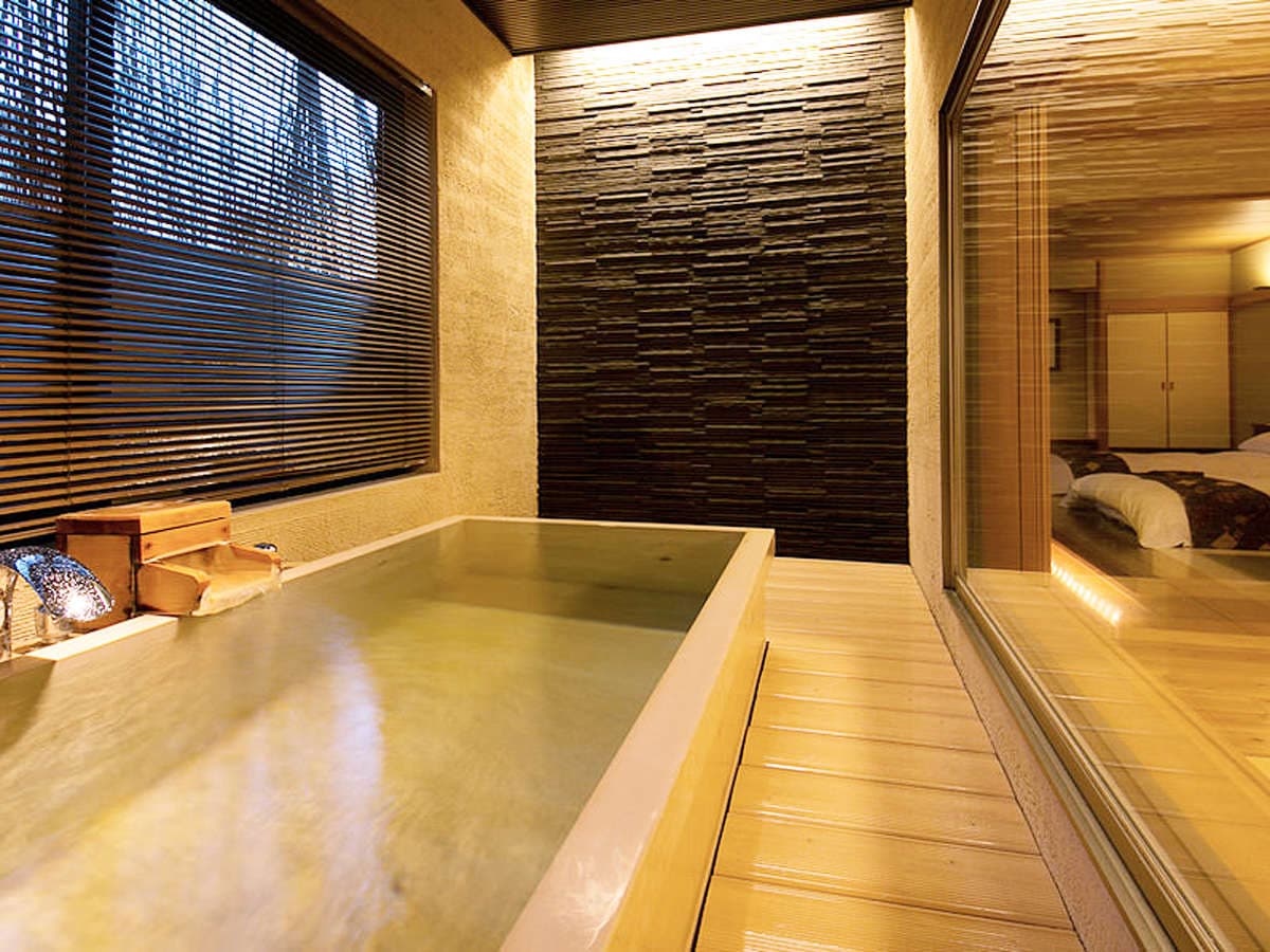 Warmly lit in-room cypress bath at Hakuba Hifumi with stacked-slate accent wall and view into the bedroom