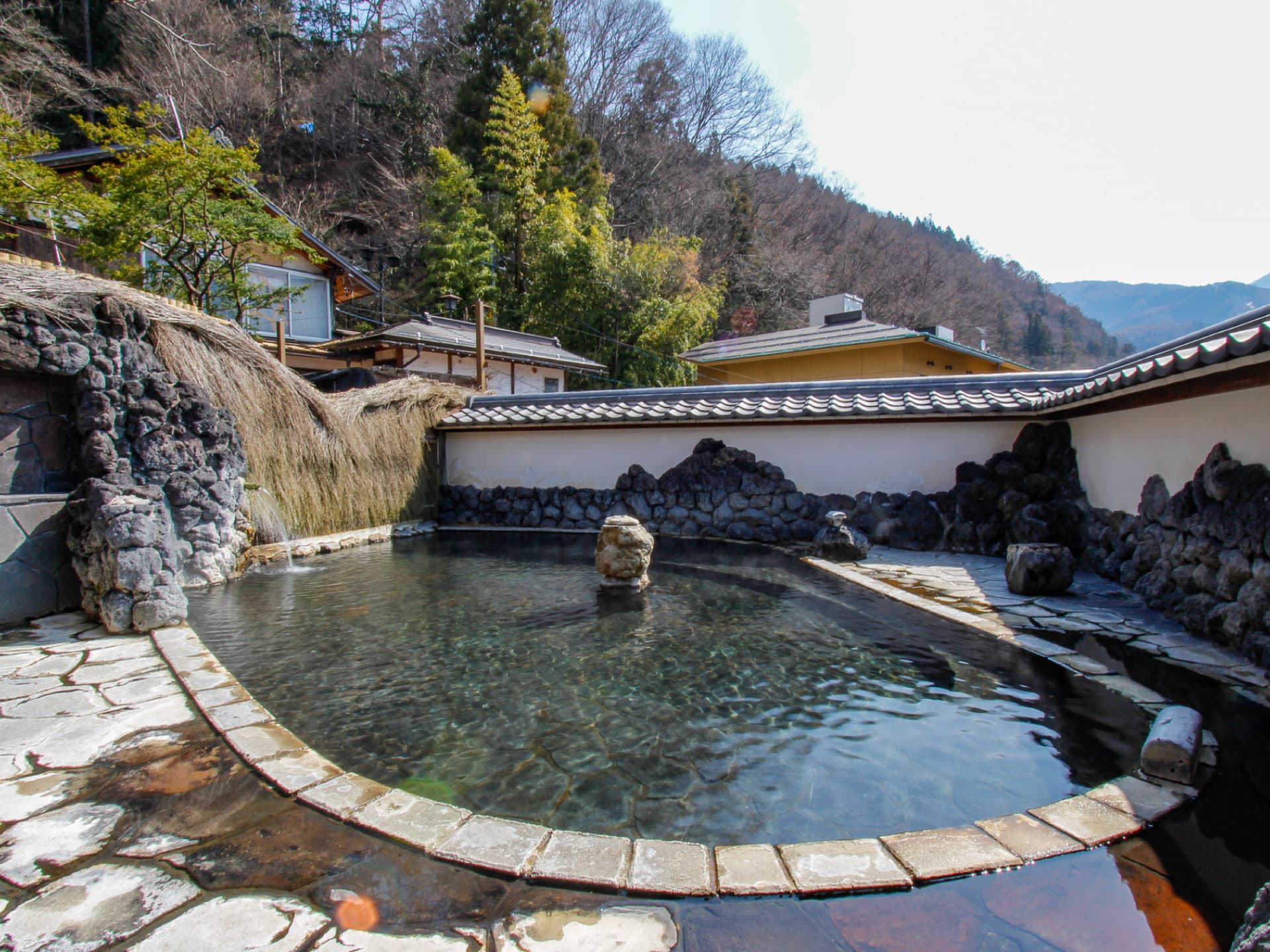 Outdoor rotenburo edged with volcanic rock under a tiled roof, forested hillside behind