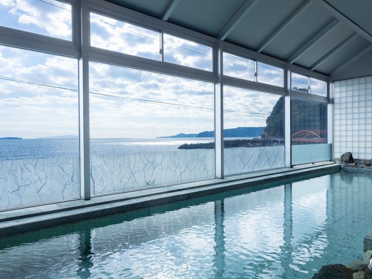Indoor onsen bath with floor-to-ceiling windows framing the Atami coastline