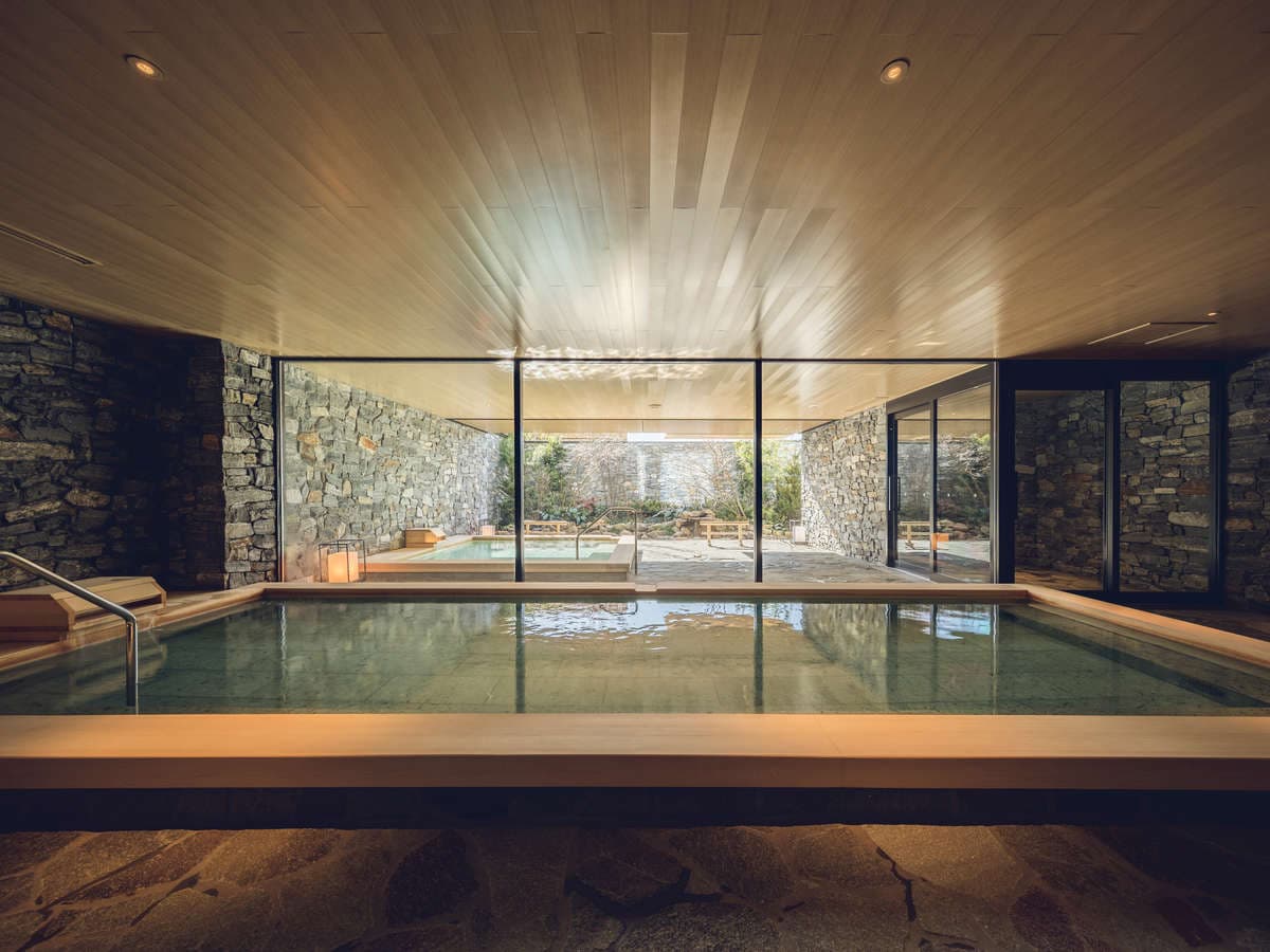 Musubi no Yu indoor onsen bath with a cypress ceiling and stone walls opening to an outdoor garden bath