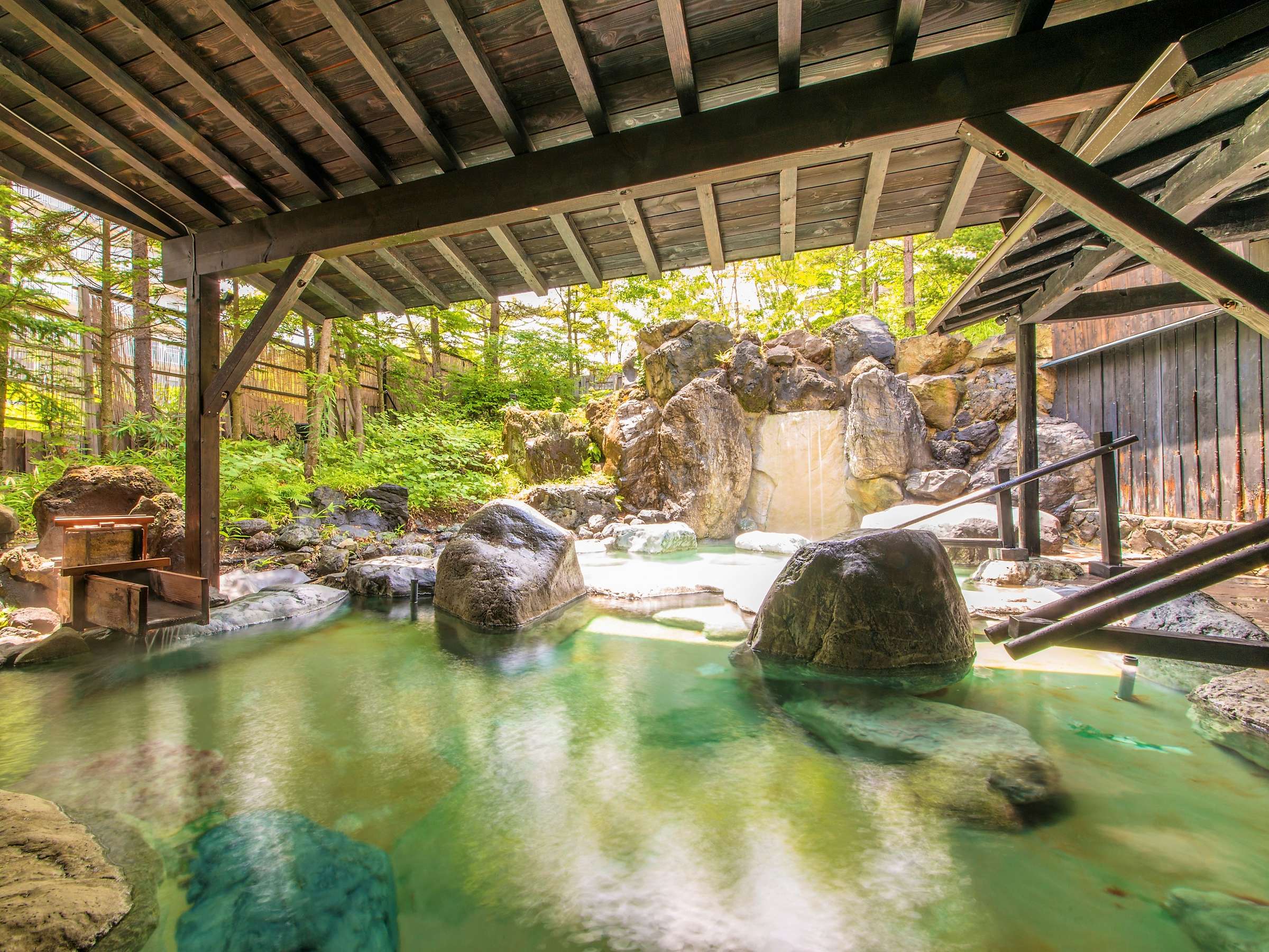 Kusatsu Now Resort Hotel open-air rotenburo onsen with large rocks, a small waterfall and green forest backdrop under a timber roof
