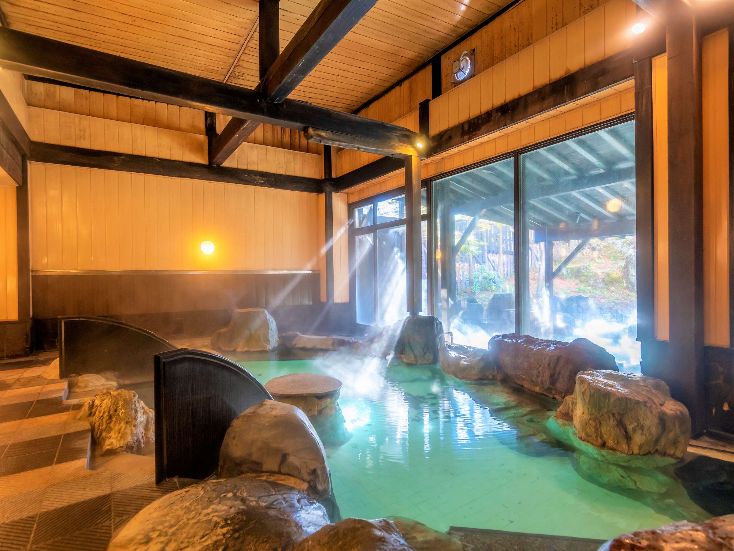 Kusatsu Now Resort Hotel indoor onsen bath with rising steam, rock-lined pool and forest view through tall windows