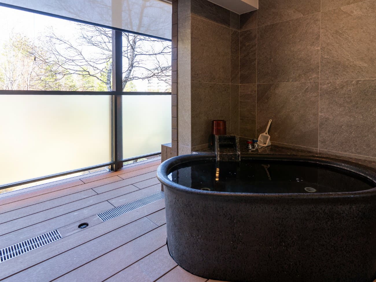 Kusatsu Now Resort Hotel private in-room onsen bath, black stone tub on a wood deck beside a frosted-glass terrace