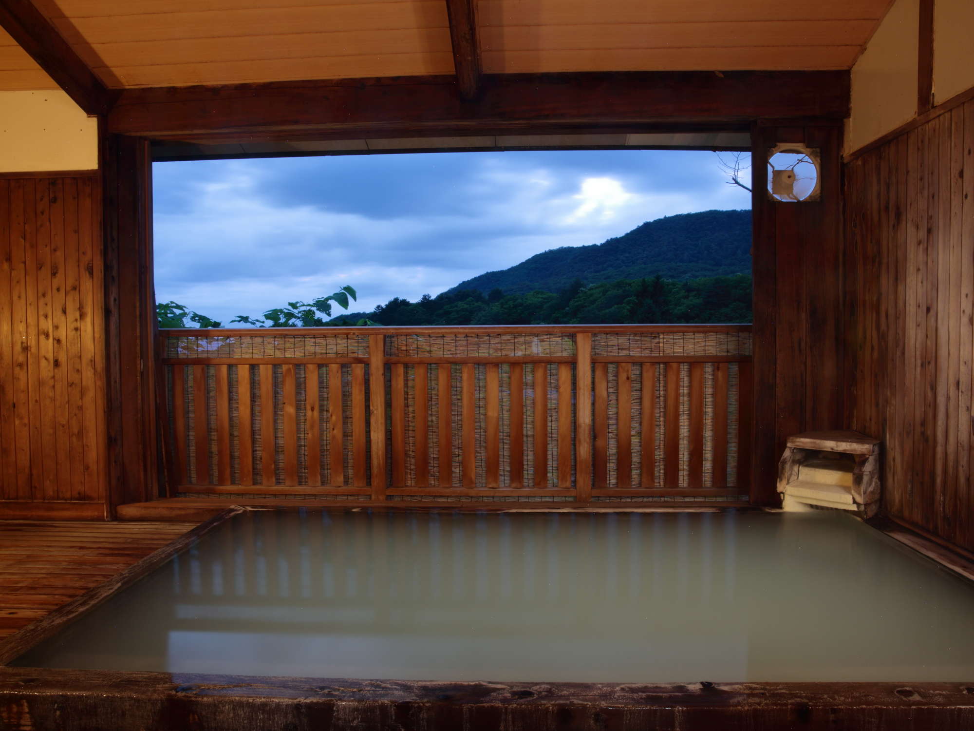 Wood-framed open-air onsen bath at dusk with a mountain view through a bamboo railing at Zao Onsen