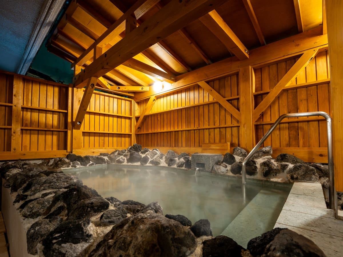 Larger outdoor rotenburo bath edged with lava rock and steam under a timber-frame pavilion