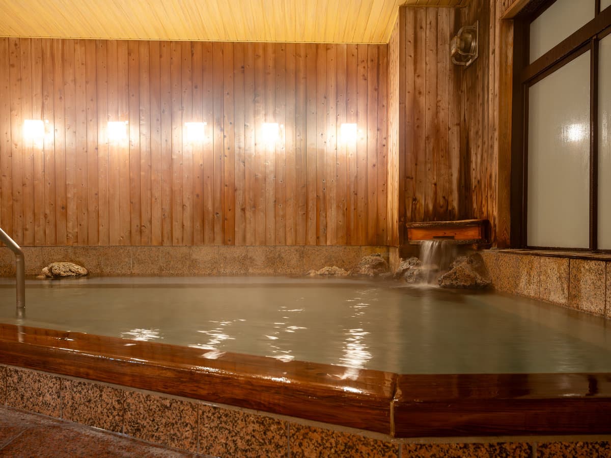 Wood-panelled indoor onsen bath with a wooden spout pouring thermal water and rising steam
