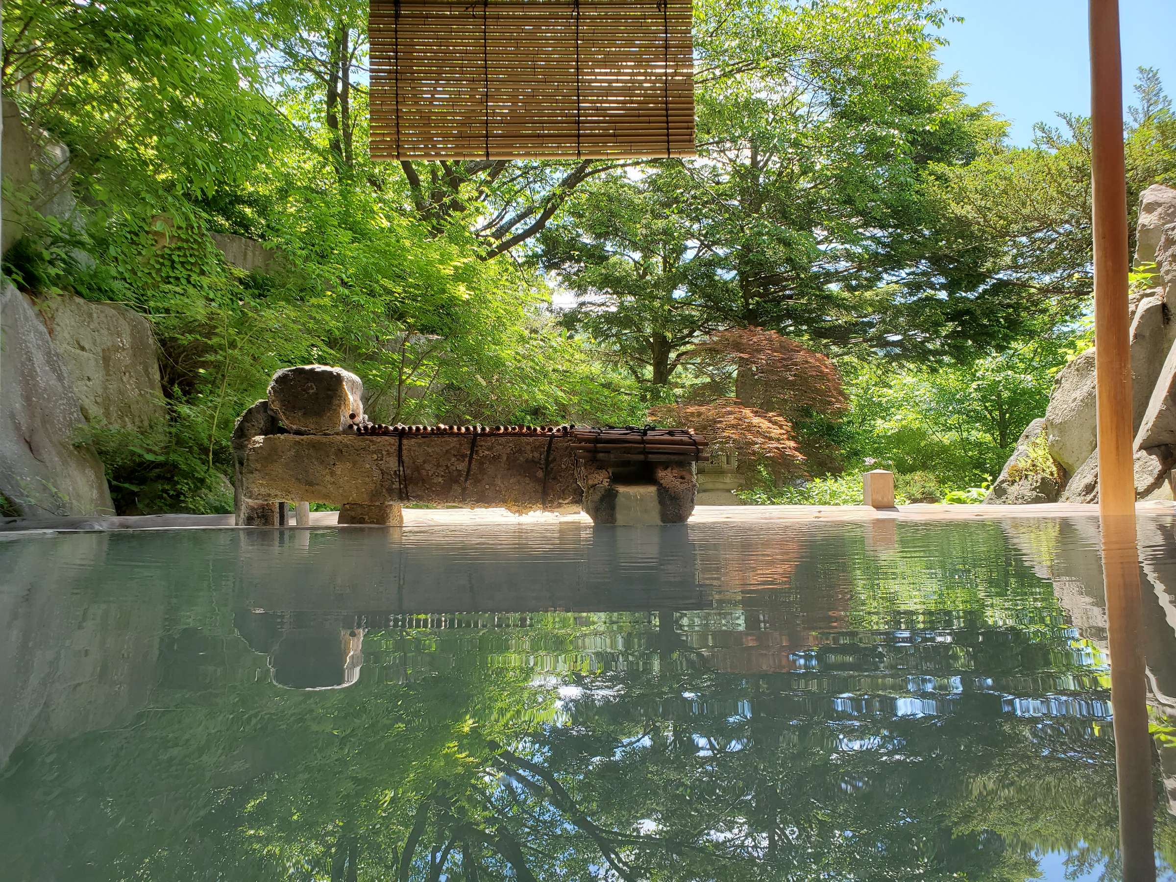 Milky sulfur open-air rotenburo bath fed by a bamboo spout amid summer greenery at Zao Plaza Hotel