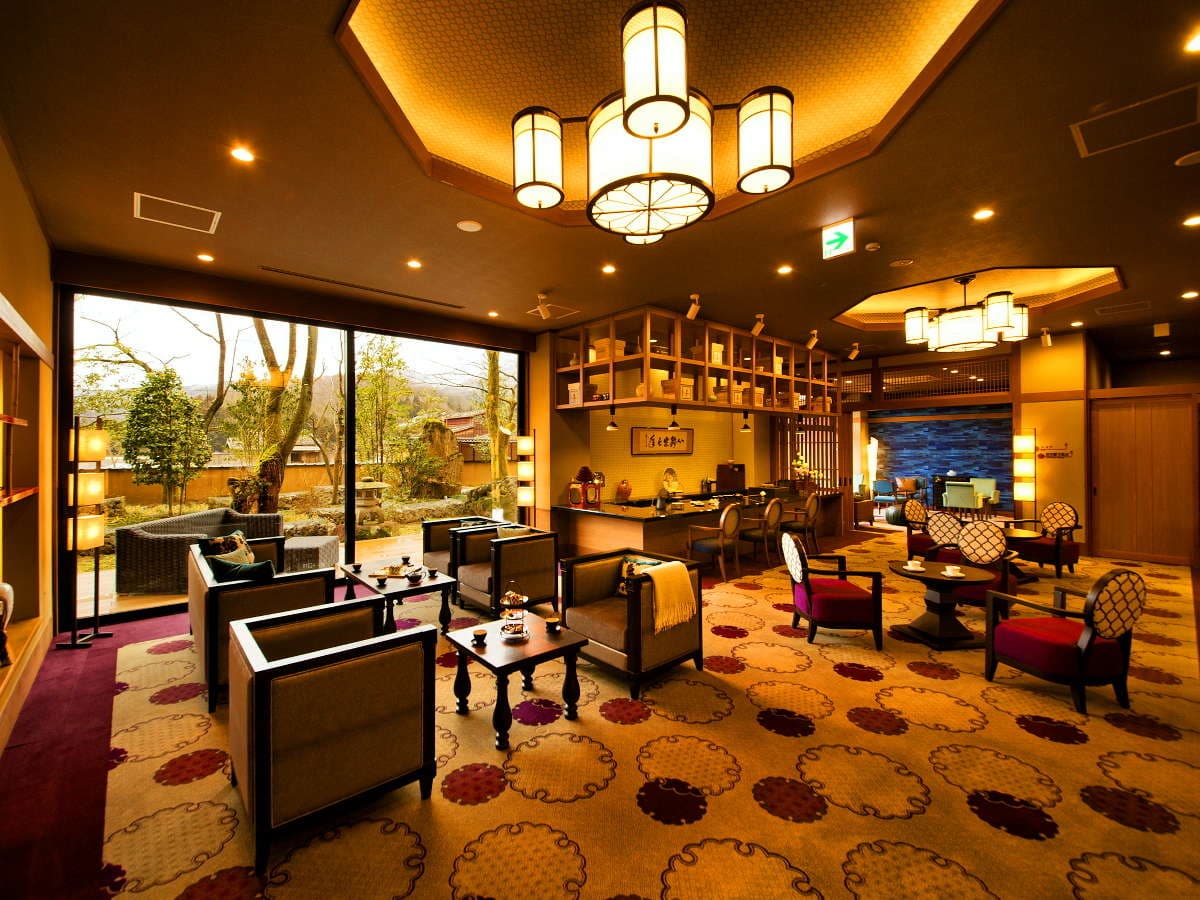 Warmly lit lobby lounge with patterned carpet, armchairs and garden-view windows at Kanazawa Yuwaku Onsen Hyakurakuso