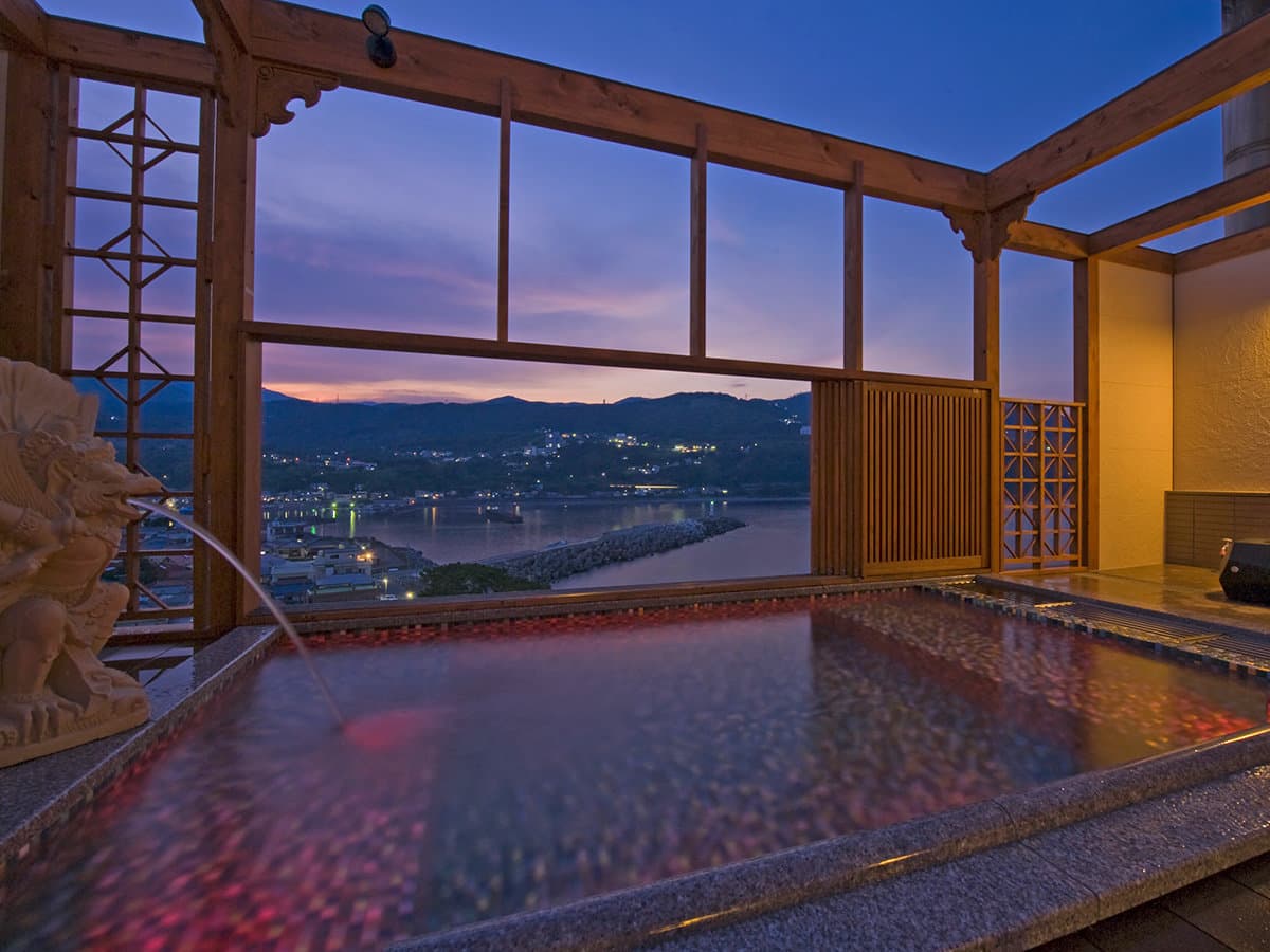 Illuminated open-air onsen bath at dusk overlooking the bay lights of Inatori at Hama no Yu