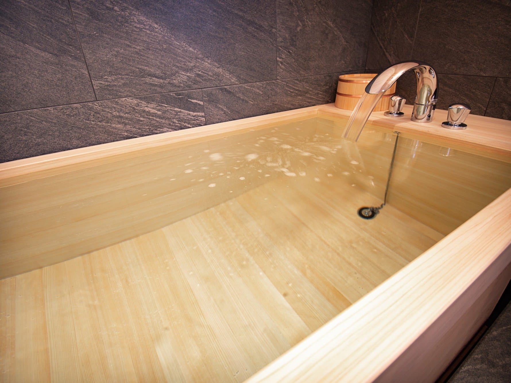 Hinoki cypress soaking bathtub filling with clear water against a dark stone wall in a private bath at Kaze no Naramachi