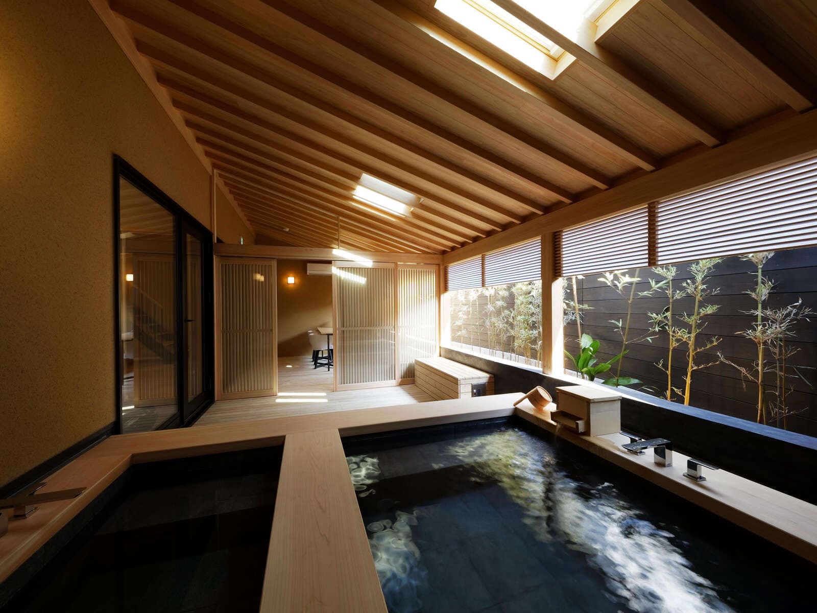 Indoor stone-and-wood hot spring bath with skylight ceiling and bamboo garden view at Koto no Niwa