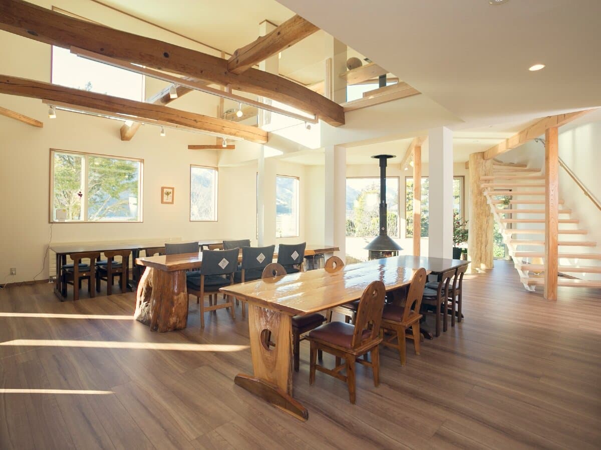 Spacious lounge and dining hall at Ryokan Mokuzin with exposed wood beams, wood-slab tables and a central wood stove