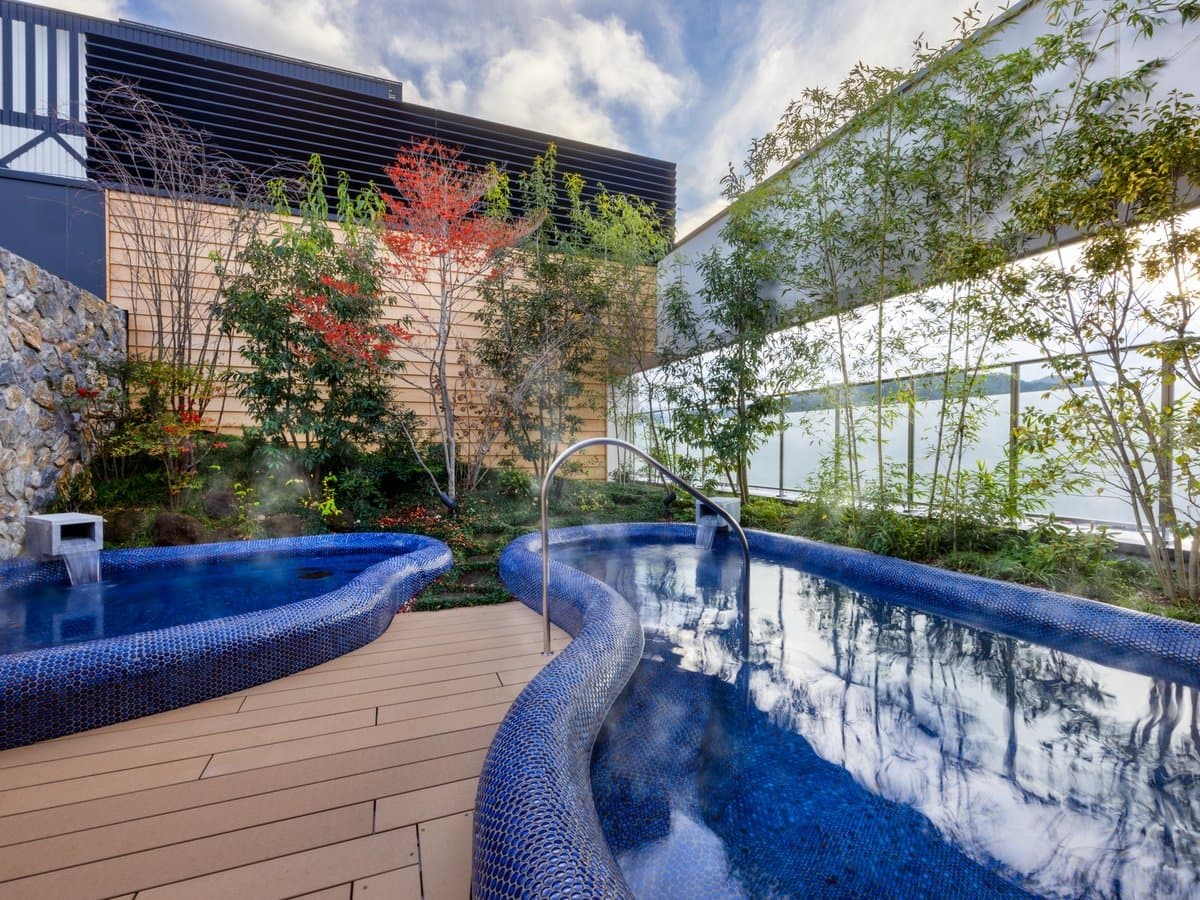 Open-air rotenburo bath with blue mosaic tiling and a sun lounger on a wooden deck beside a planted garden