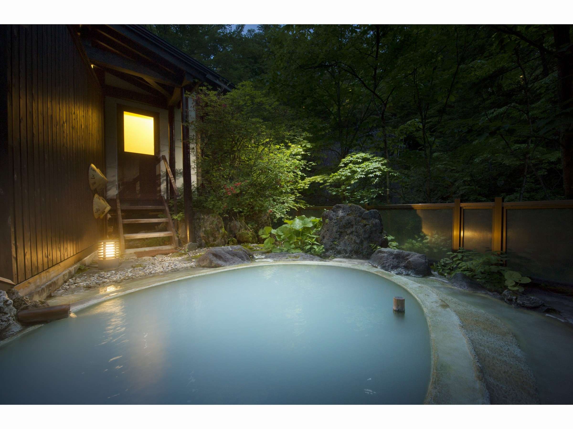 Milky-white outdoor rotenburo bath at Sansuikan Yugawaso beside a wooden lodge with a lit window in twilight greenery