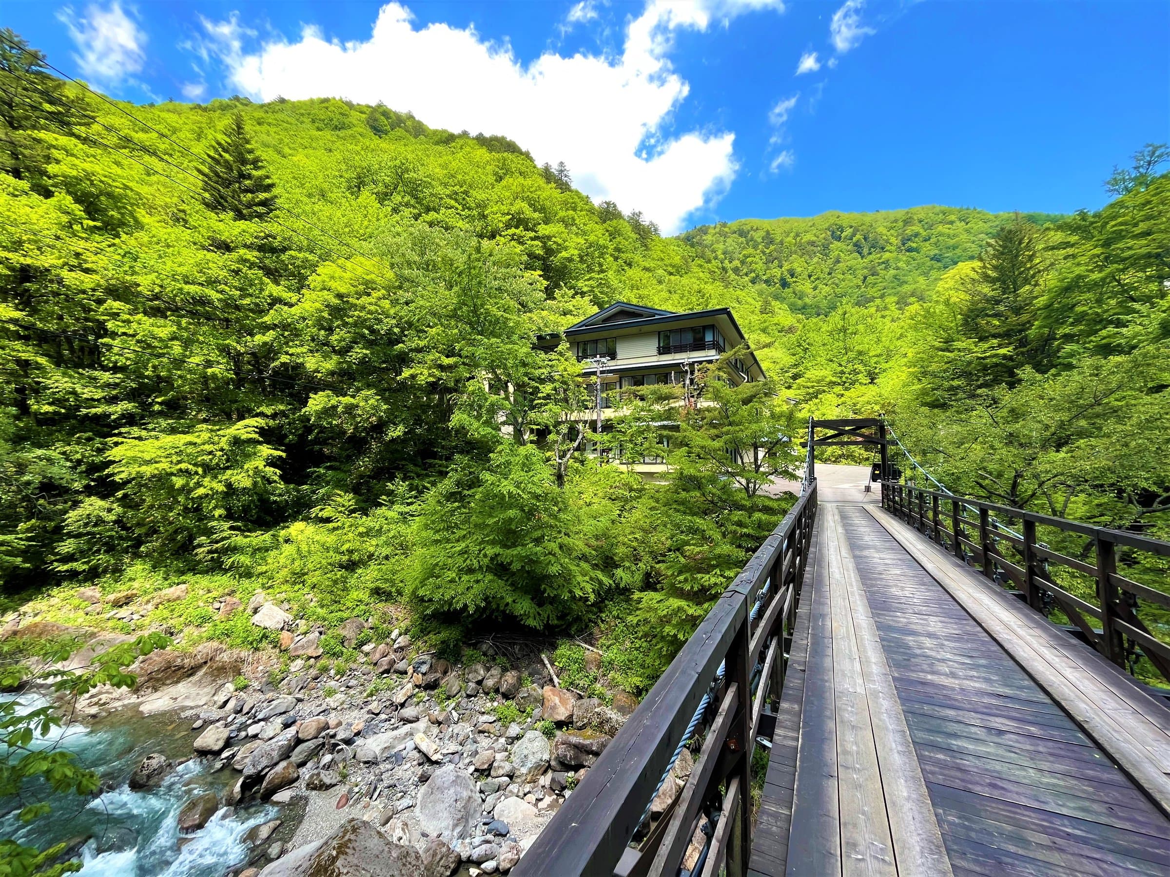 Sansuikan Yugawaso ryokan in Shirahone Onsen seen from a wooden suspension bridge over a mountain stream in lush summer greenery