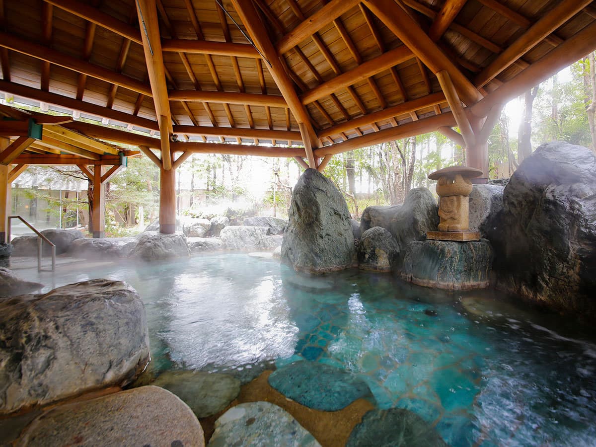 Open-air rotenburo onsen with steaming water, natural rocks and a wooden pavilion at Takayama Green Hotel