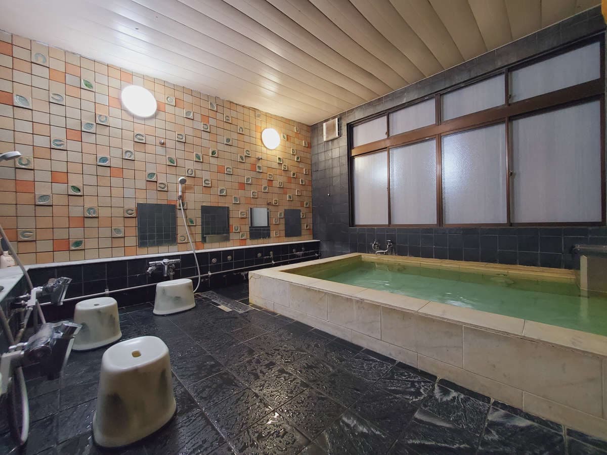 Retro mosaic-tiled public bath with a stone soaking tub of green onsen water at Ryokan Murayama