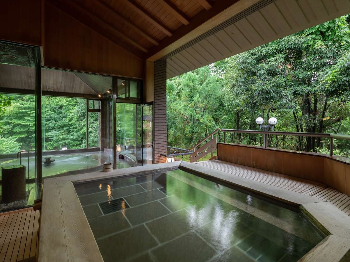 Open-air stone rotenburo with a wooden deck and steps overlooking lush green forest at Yunode Ryokan, Yuwaku Onsen