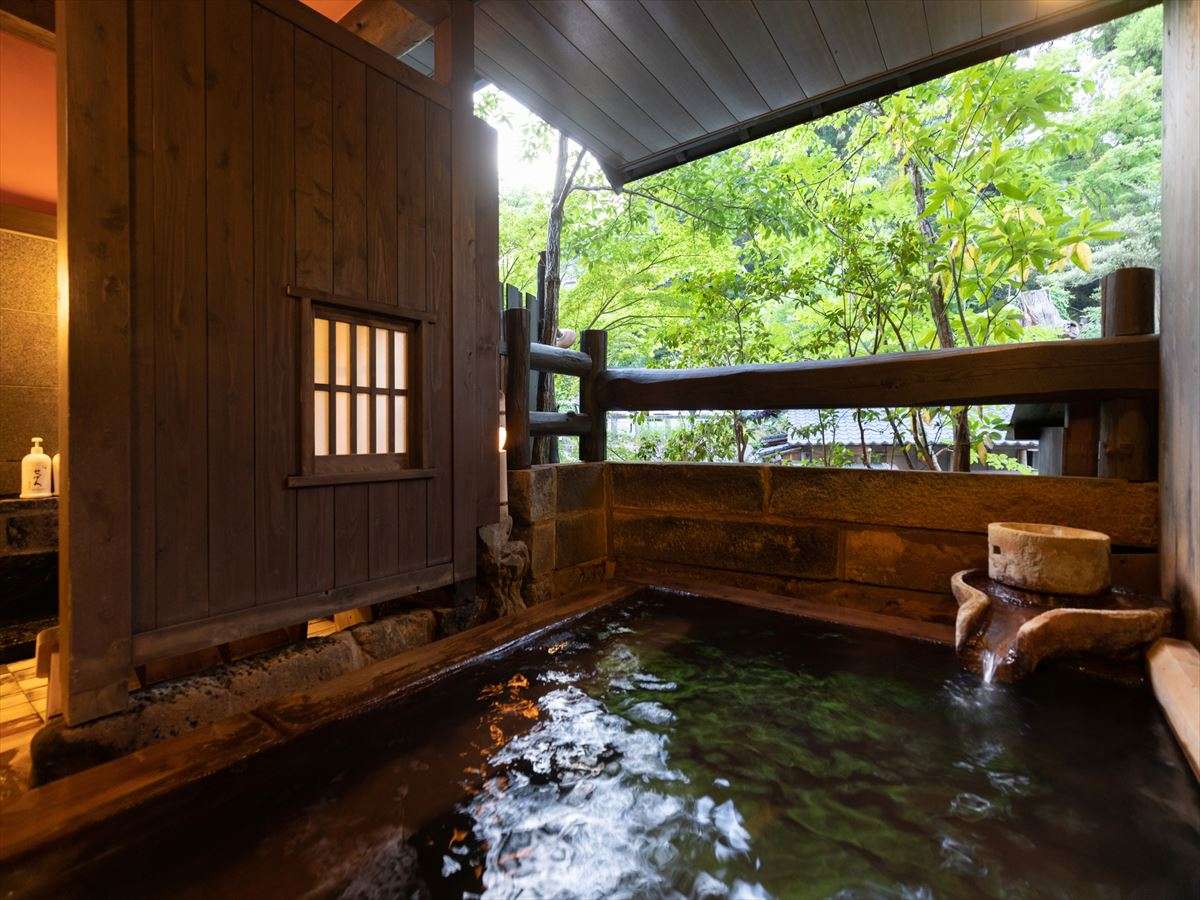Open-air rotenburo bath at Fujiya surrounded by fresh greenery, with a stone water spout and timber railing