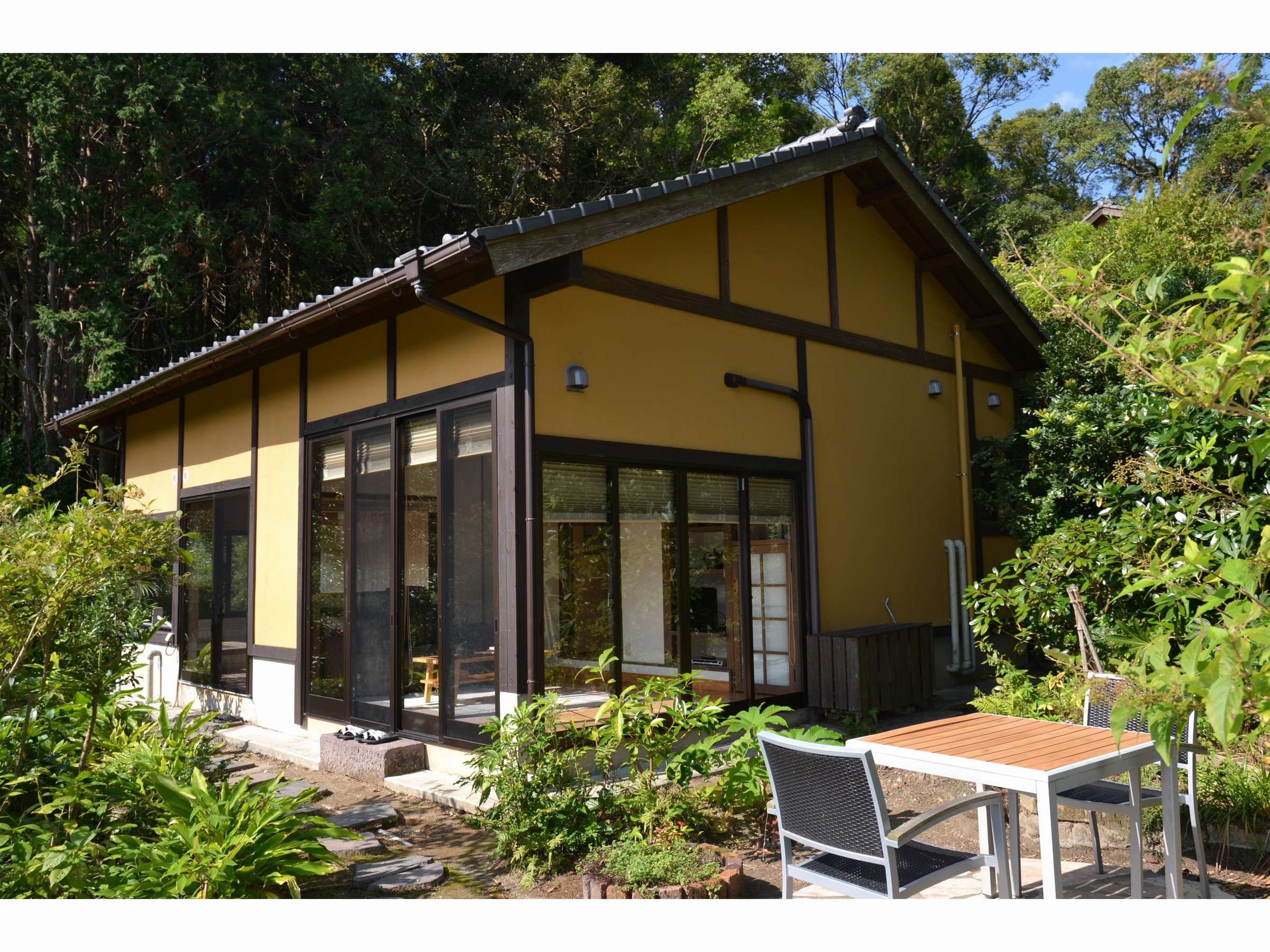 Exterior of a detached ochre-walled villa at Yurian with private terrace and dining set framed by forest, Ibusuki