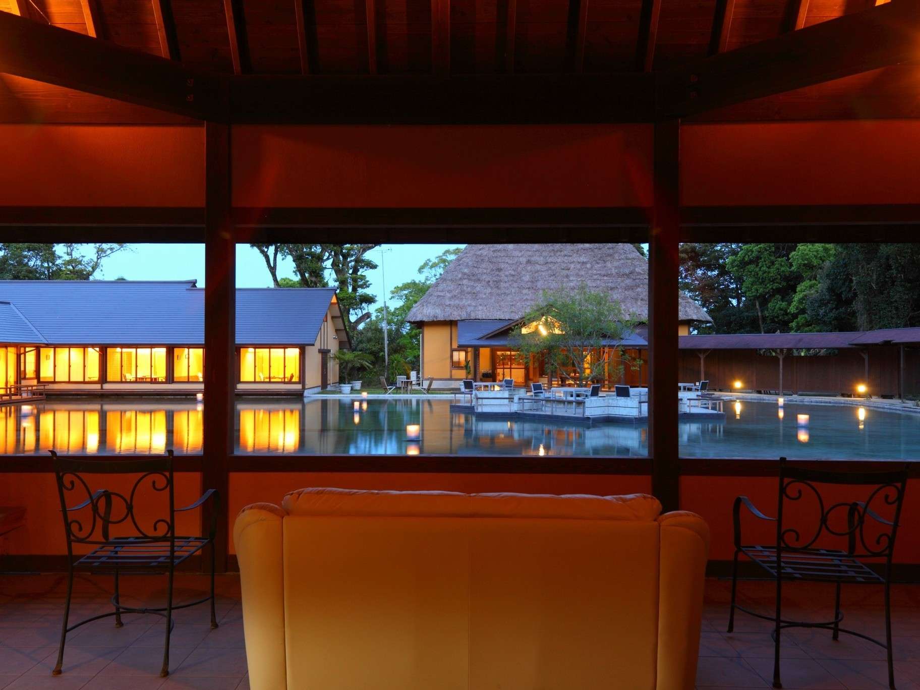 Yurian lounge interior at dusk looking out over the lantern-lit reflecting pond and thatched buildings, Ibusuki Onsen