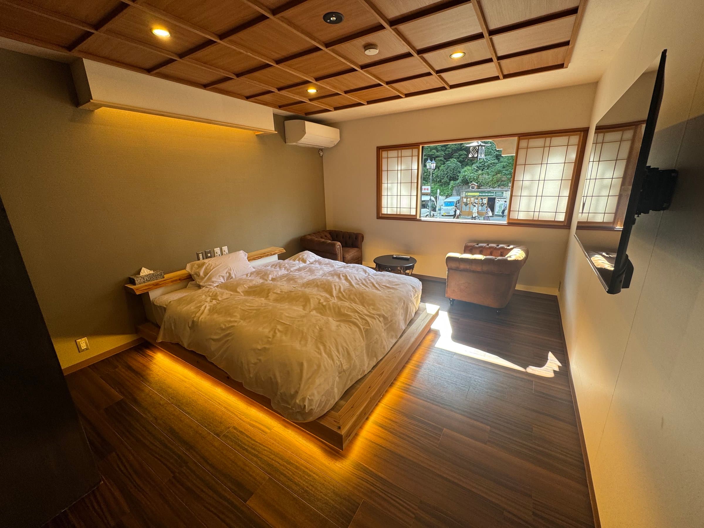 Modern guest room with low platform bed, indirect lighting and leather armchairs facing a street-view window at Yanadaya