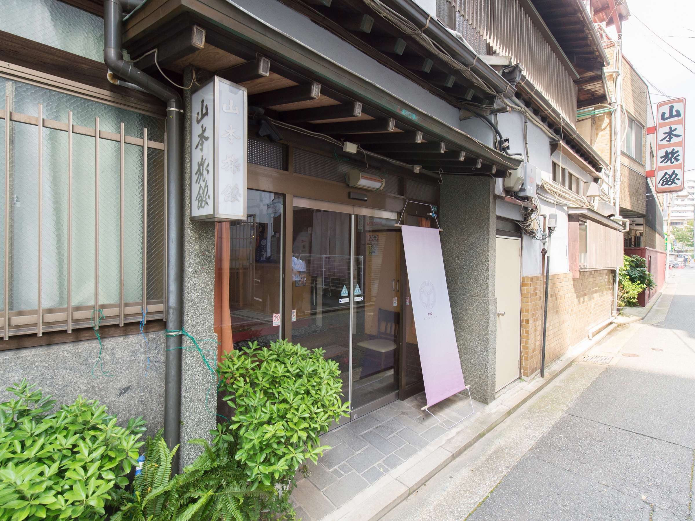 Quiet street-side entrance approach to Yamamoto Ryokan with greenery, a noren-style banner and vertical name lanterns
