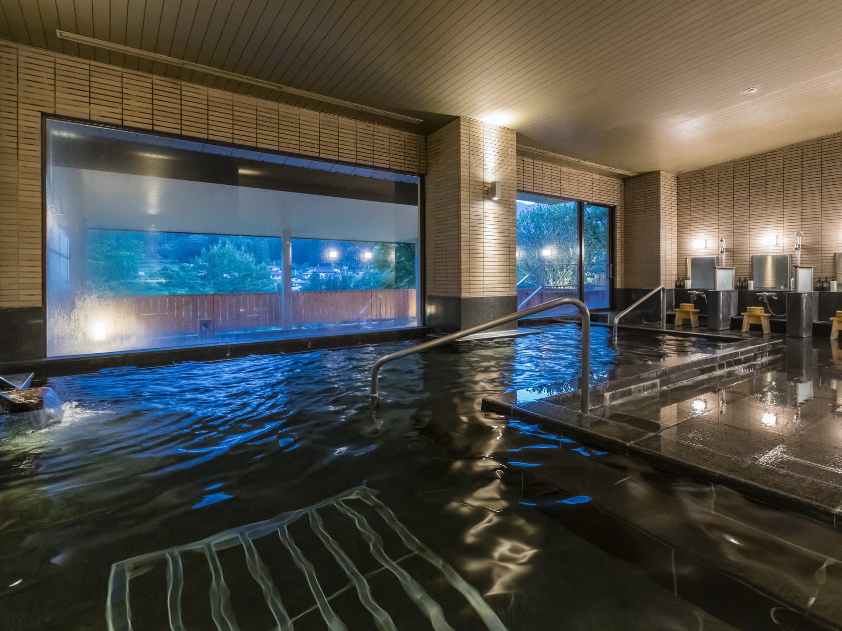 Large indoor onsen bath with stone surround and forest-view window lit in the evening at Wakatake no Sho, Kinugawa Onsen