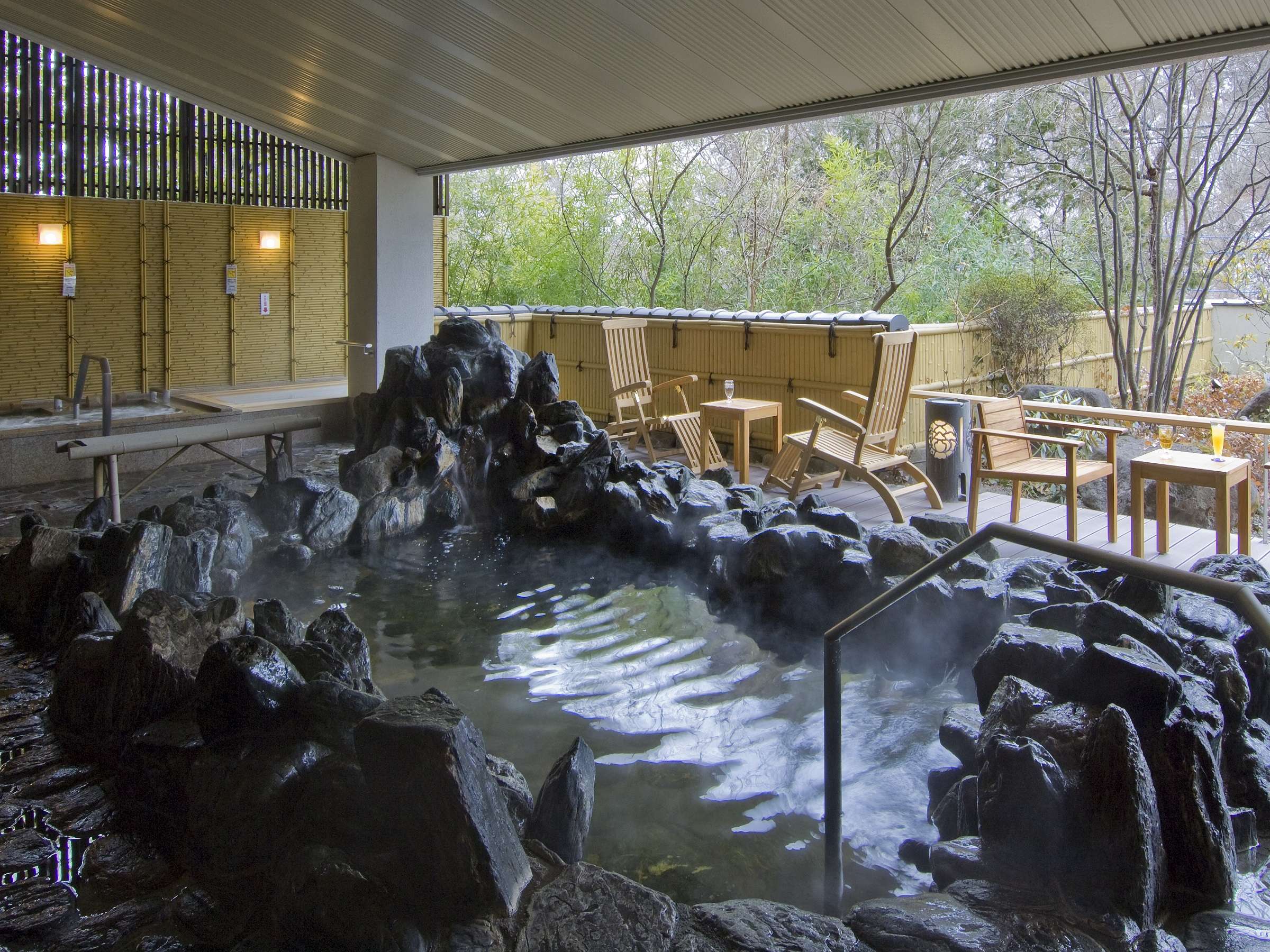 Steaming rocky outdoor onsen bath under a covered deck overlooking woodland at Nikko Senhime Monogatari