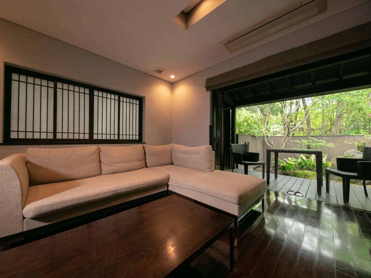 Guest room lounge with a sofa opening onto a wooden terrace and green garden at Yufuin Bettei Itsuki