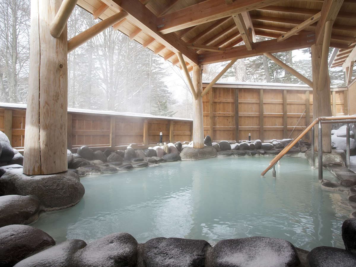 Snow-covered outdoor rotenburo with steaming milky hot spring water and a log pavilion in winter at Okunikko Kogen Hotel