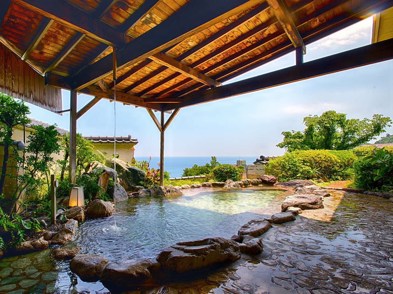 Open-air rock rotenburo onsen under a timber pavilion with greenery and distant ocean view at Atagawa Prince Hotel