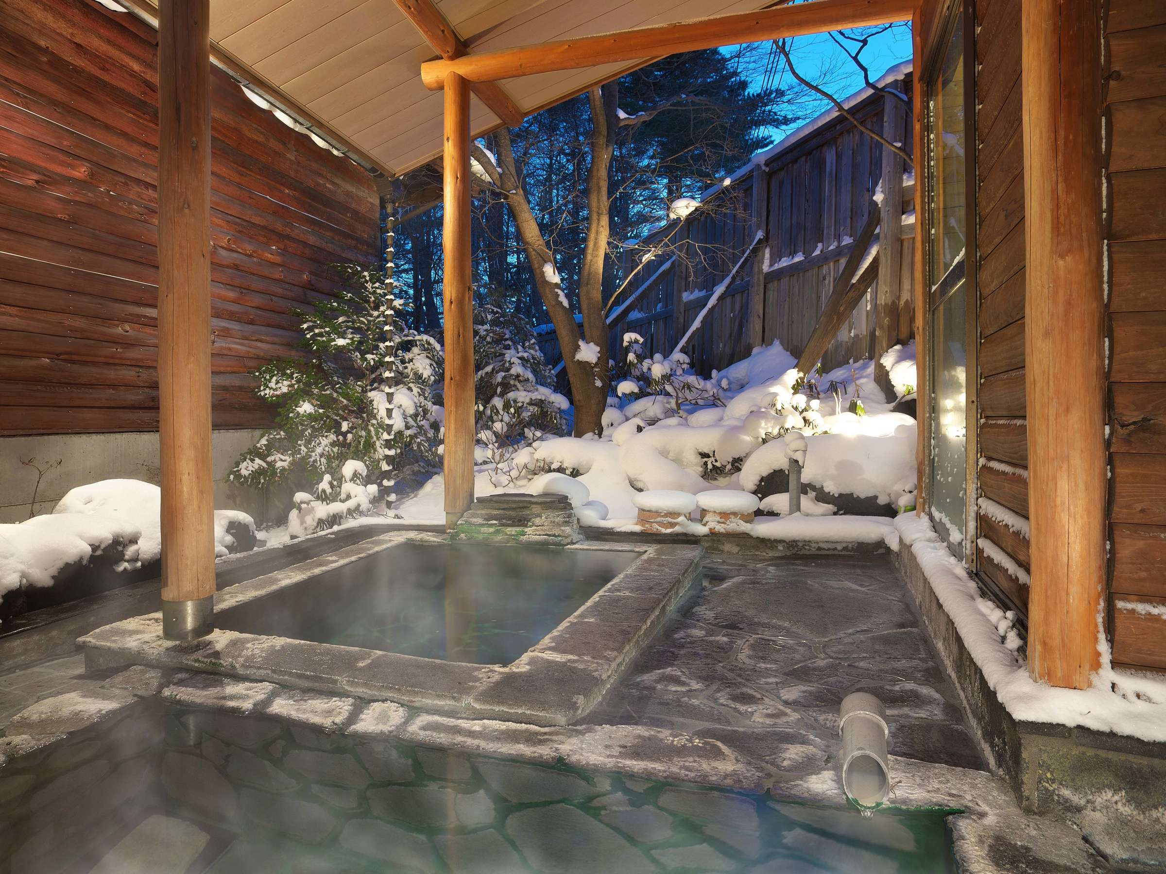 Stone outdoor hot-spring bath steaming under a wooden eave surrounded by snow-covered garden