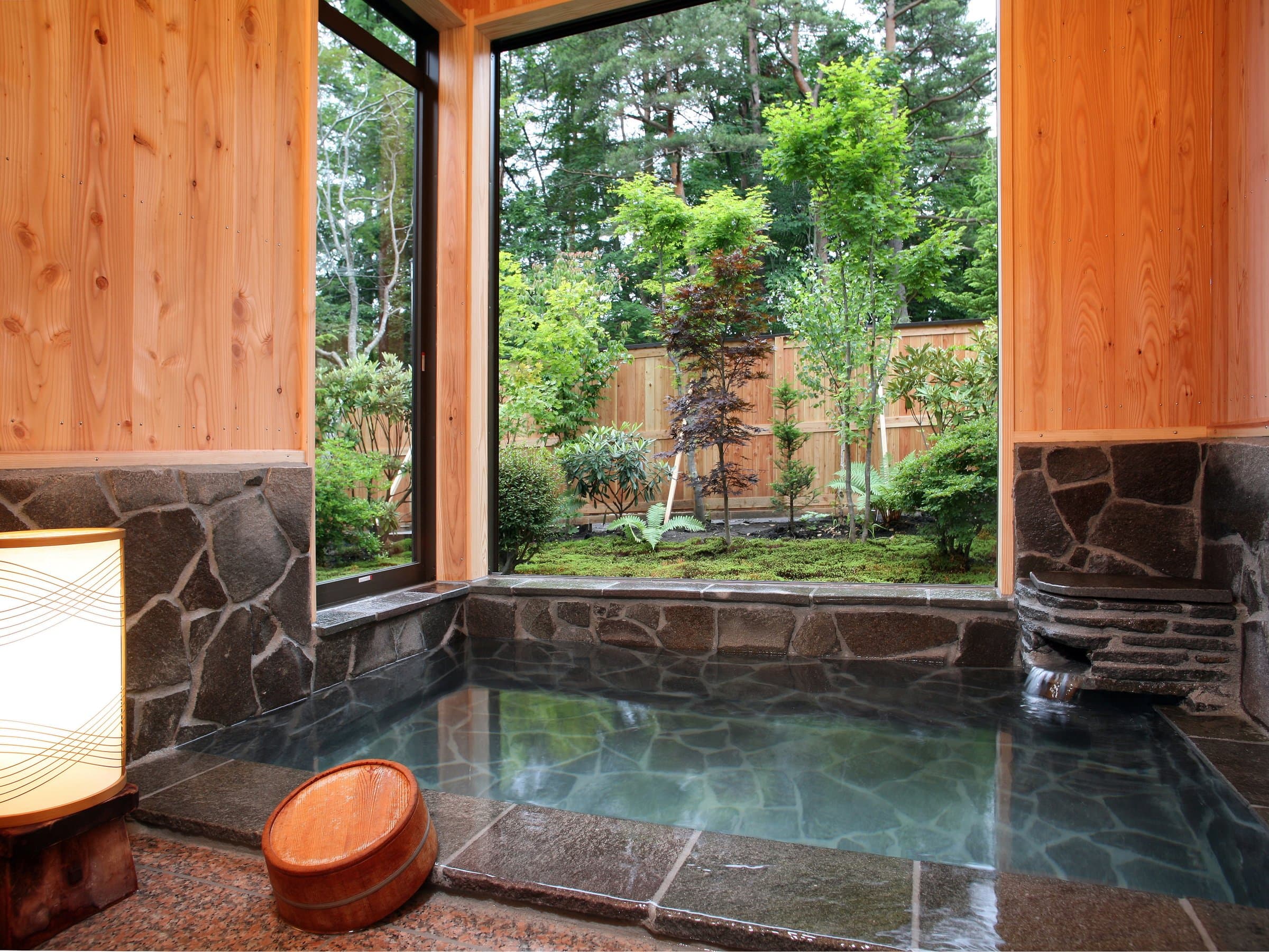 Indoor stone hot-spring bath with a picture window overlooking a green Japanese garden