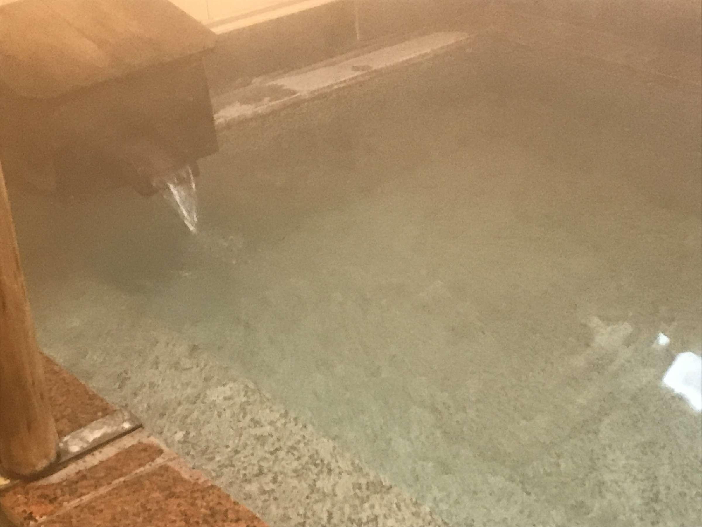 Close view of milky Kusatsu hot spring water flowing from a wooden spout into a tiled bath with rising steam at Suehiroya Ryokan
